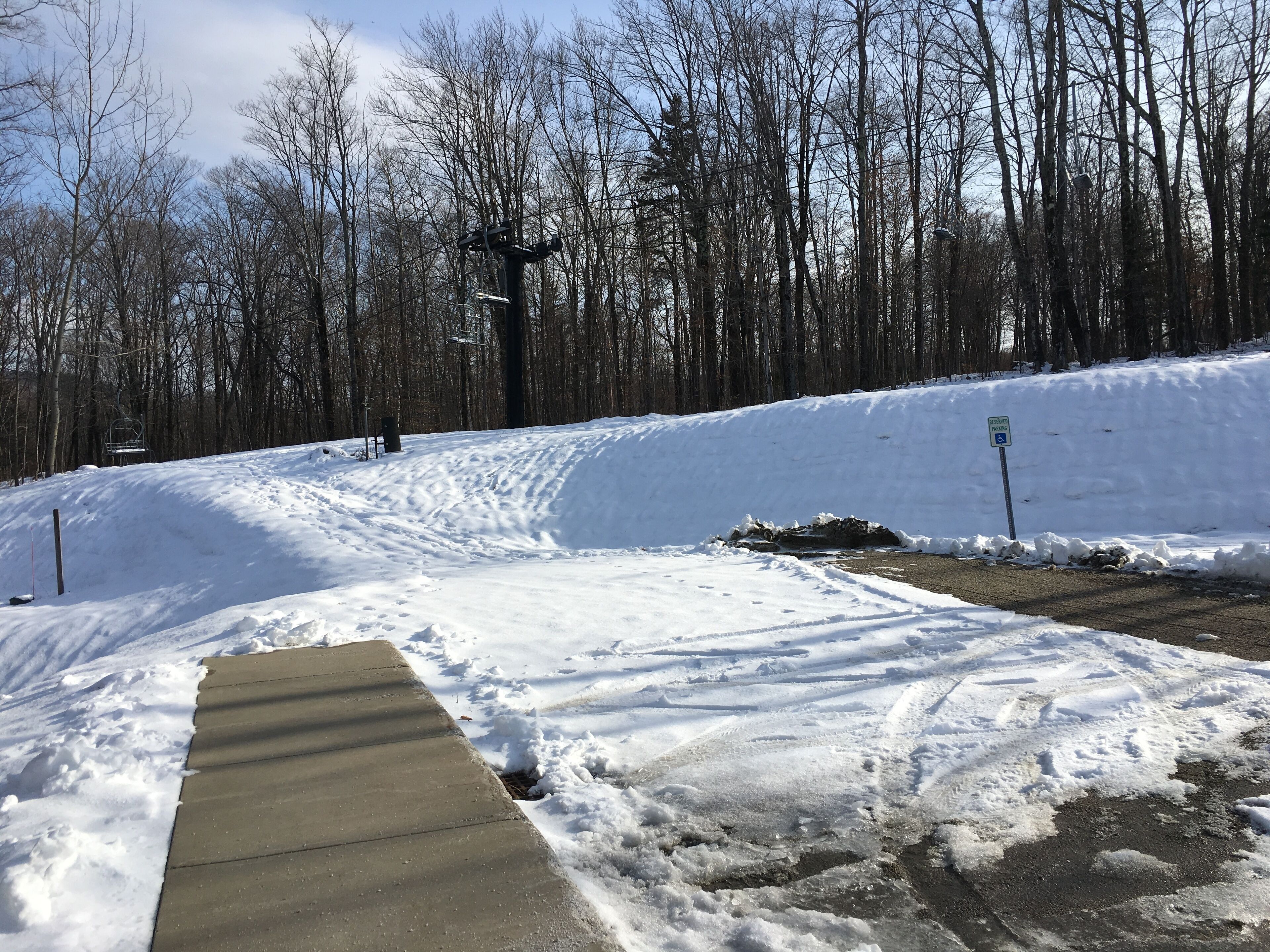 A snow-covered parking area with a concrete ramp leading to a parking lot, surrounded by trees.