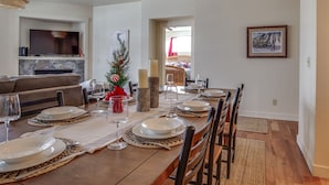 Un comedor con una mesa de madera dispuesta para seis personas, rodeada de sillas. Un televisor está montado sobre la chimenea, y se ve un árbol de Navidad al fondo.