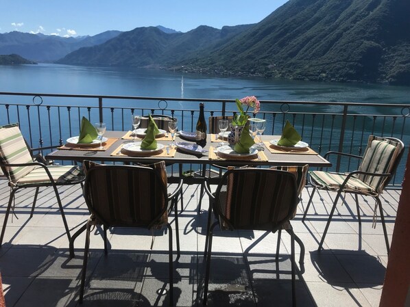 A dining table set with plates, glasses, and a bottle, surrounded by chairs, overlooking a lake with mountains in the background.