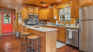 A wooden cabin interior with a kitchen featuring wooden cabinets, a stainless steel refrigerator, and a bar area with stools. - Creekside Serenade relax by the running creek (Broken Bow)