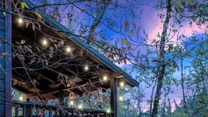 A wooden cabin with a balcony, string lights, and a fire pit area with red Adirondack chairs. - Creekside Serenade relax by the running creek (Broken Bow)