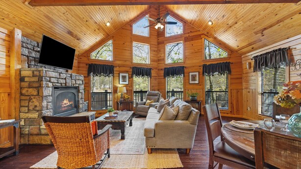 A cozy log cabin interior with a stone fireplace, a television mounted above it, a comfortable sofa, and a dining table set for four. - Creekside Serenade relax by the running creek (Broken Bow)
