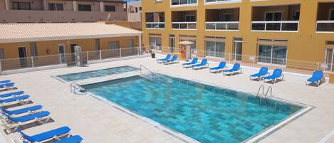 Une piscine sur le toit, avec des chaises longues bleues, et un bâtiment jaune doté de balcons.