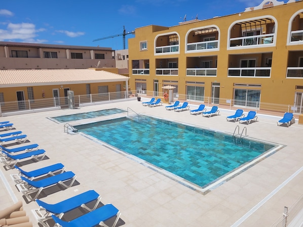 Una piscina en la azotea con sillas reclinables azules y un edificio amarillo con balcones.