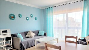 Una sala de estar con un sofá gris, una mesa de centro blanca y sillas de madera. Hay un mueble blanco para televisión con un microondas y libros. La habitación tiene paredes azul claro y un ventanal con cortinas.