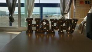 Una habitación con vista a la playa, una mesa con letras de madera que dicen 'Happy' y un estante de metal con objetos coloridos.