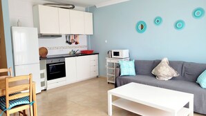 Cocina moderna con armarios blancos, un microondas y una cafetera sobre la encimera. En la sala de estar, hay un sofá gris con un cojín floral y una mesa de centro blanca.