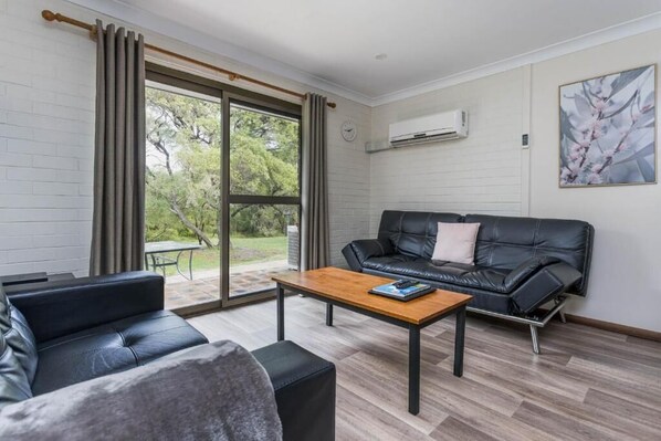 A modern living room with a black leather sofa, a wooden coffee table, and a large window with curtains.