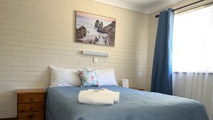 Une chambre à coucher avec un lit, des oreillers, une table de chevet, un tableau et une fenêtre avec des rideaux.