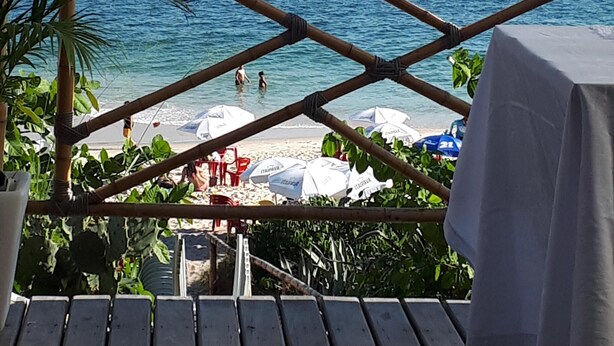 Une terrasse en bois donnant sur une plage, avec des parasols et des personnes se baignant.