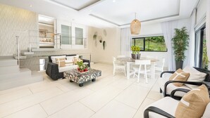 A modern living room with a sofa, coffee table, and dining area. There is a staircase leading to an upper level. - 5 star BEACH SIDE LUXURY APARTMENT/CHEF SERVICE BKFAST (Las Terrenas)