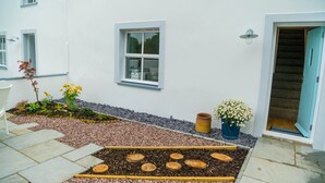 A white building with a garden area featuring a raised wooden bed, potted plants, and a stone pathway.