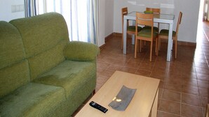 Ein Wohnzimmer mit einem grünen Sofa, einem Esstisch mit Stühlen und einer Fernbedienung auf einem Couchtisch.