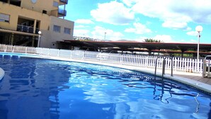 Ein Schwimmbecken mit weißem Zaun, ein Gebäude mit Balkonen und ein klarer Himmel.