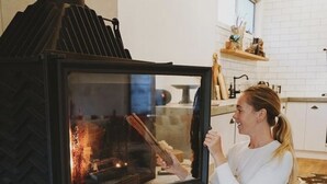 Nainen istuu keittiössä puuhellan vieressä. Keittiössä on valkoiset laatat ja kivinen takka.