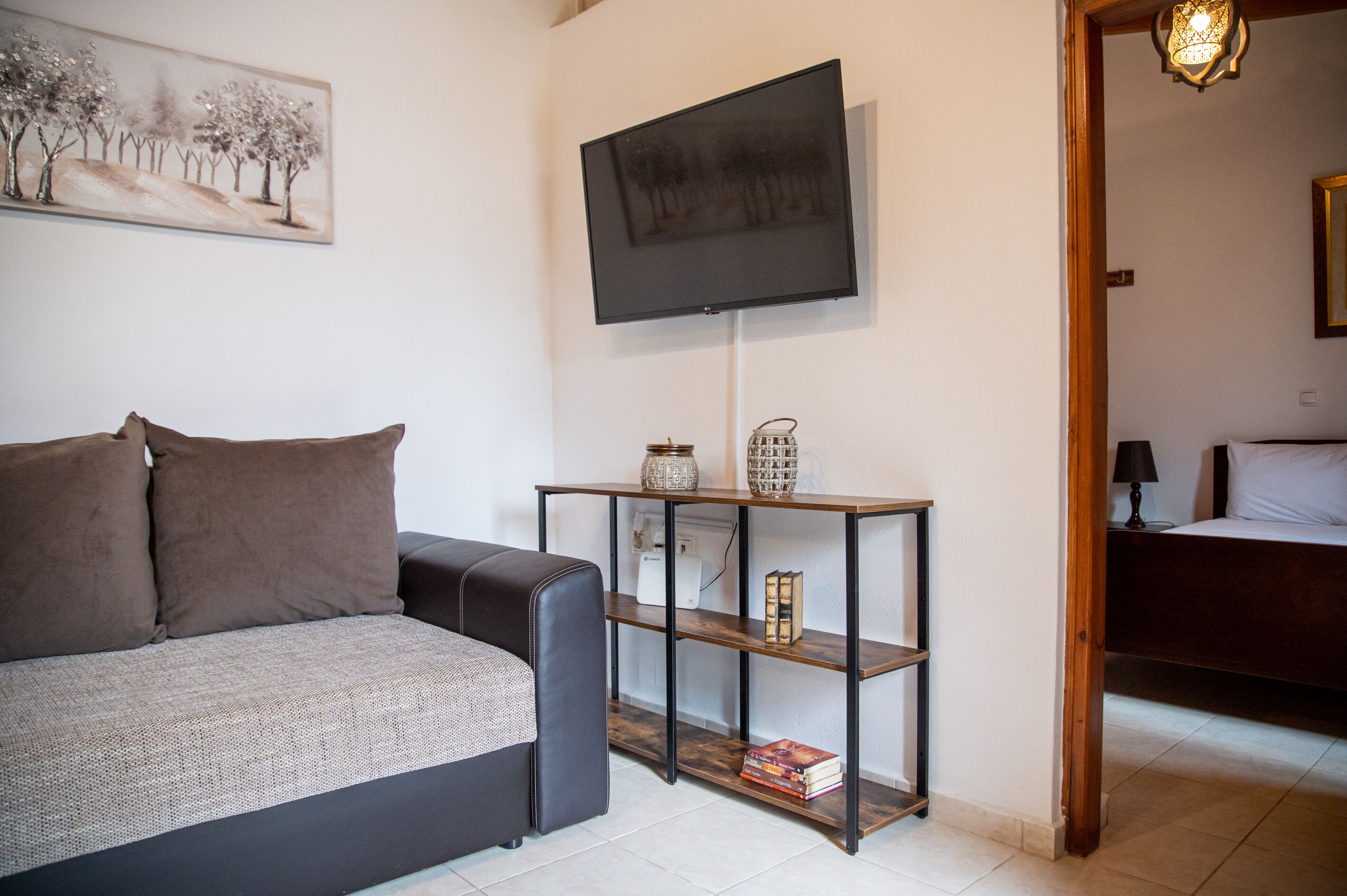 Un salon moderne avec un canapé, un téléviseur fixé au mur, une console en bois agrémentée d’objets décoratifs et un lit dans la pièce adjacente.