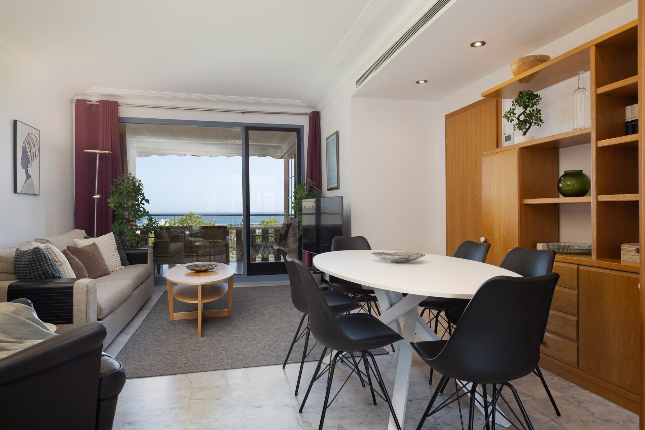 A modern living room with a sofa, coffee table, and dining area with chairs. There is a balcony with a view.