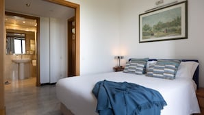 Une chambre d’hôtel avec un grand lit, une table de chevet avec une lampe, un tableau au mur et une salle de bain visible à travers une porte ouverte.