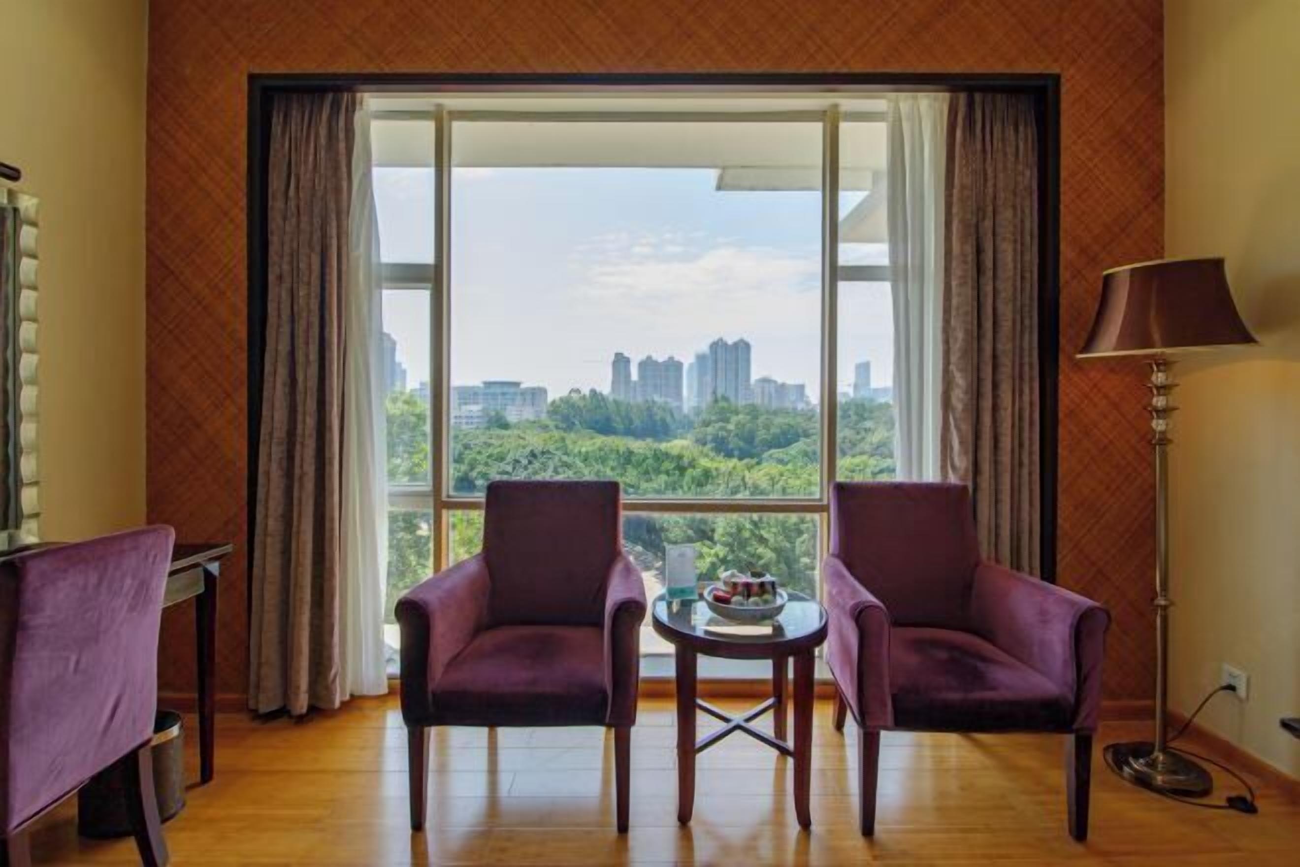 Una habitación con dos sillones morados, una pequeña mesa redonda y vistas a un paisaje urbano a través de amplios ventanales.