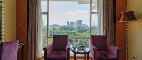 Una habitación con dos sillones morados, una pequeña mesa redonda y vistas a un paisaje urbano a través de amplios ventanales.