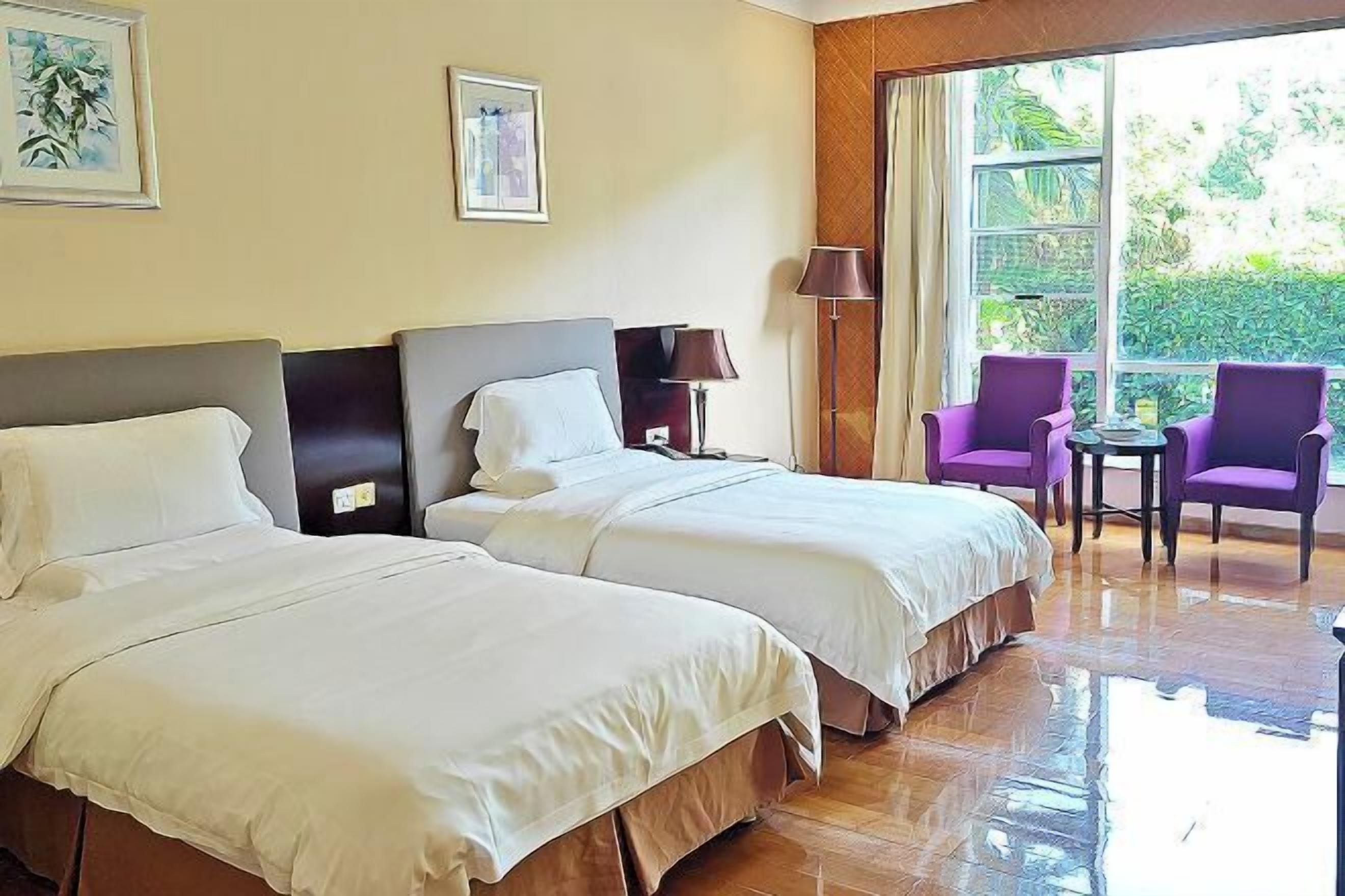 A hotel room with two beds, a balcony with purple chairs, and a large window with a view of greenery.