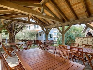An outdoor wooden pergola with tables and chairs, surrounded by a stone wall and greenery.