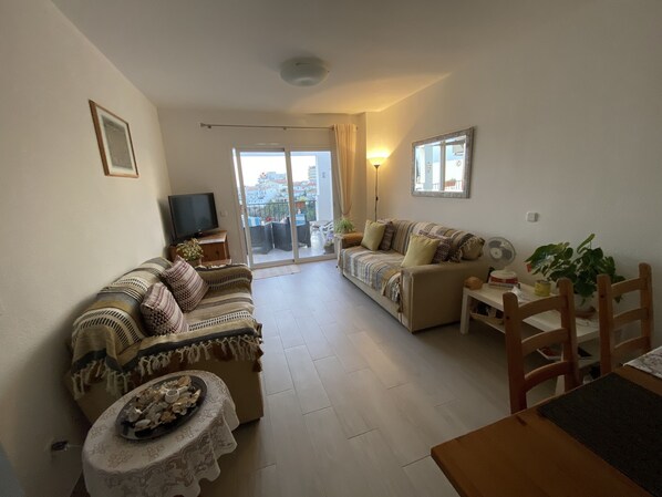 A living room with a sofa, a table, and a TV. There is a balcony with a view of buildings.