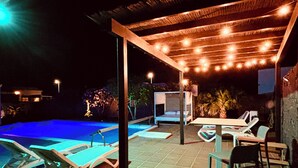Ein Poolbereich mit Liegestühlen, einem Tisch und einer Pergola mit Lichterketten.