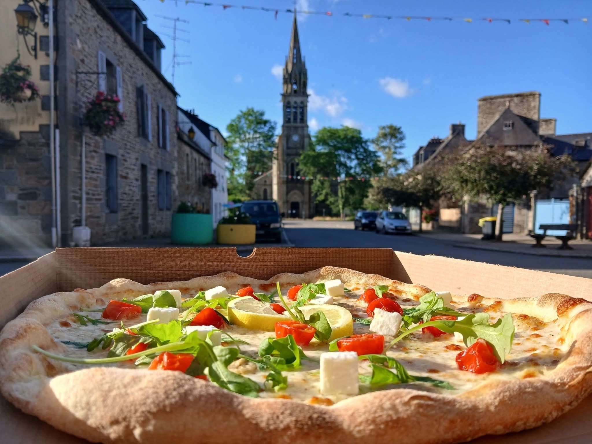 Eine Pizzaschachtel mit einer Pizza, belegt mit Salat, Tomaten und Käse, vor einer Straße mit Gebäuden und einer Kirche im Hintergrund.