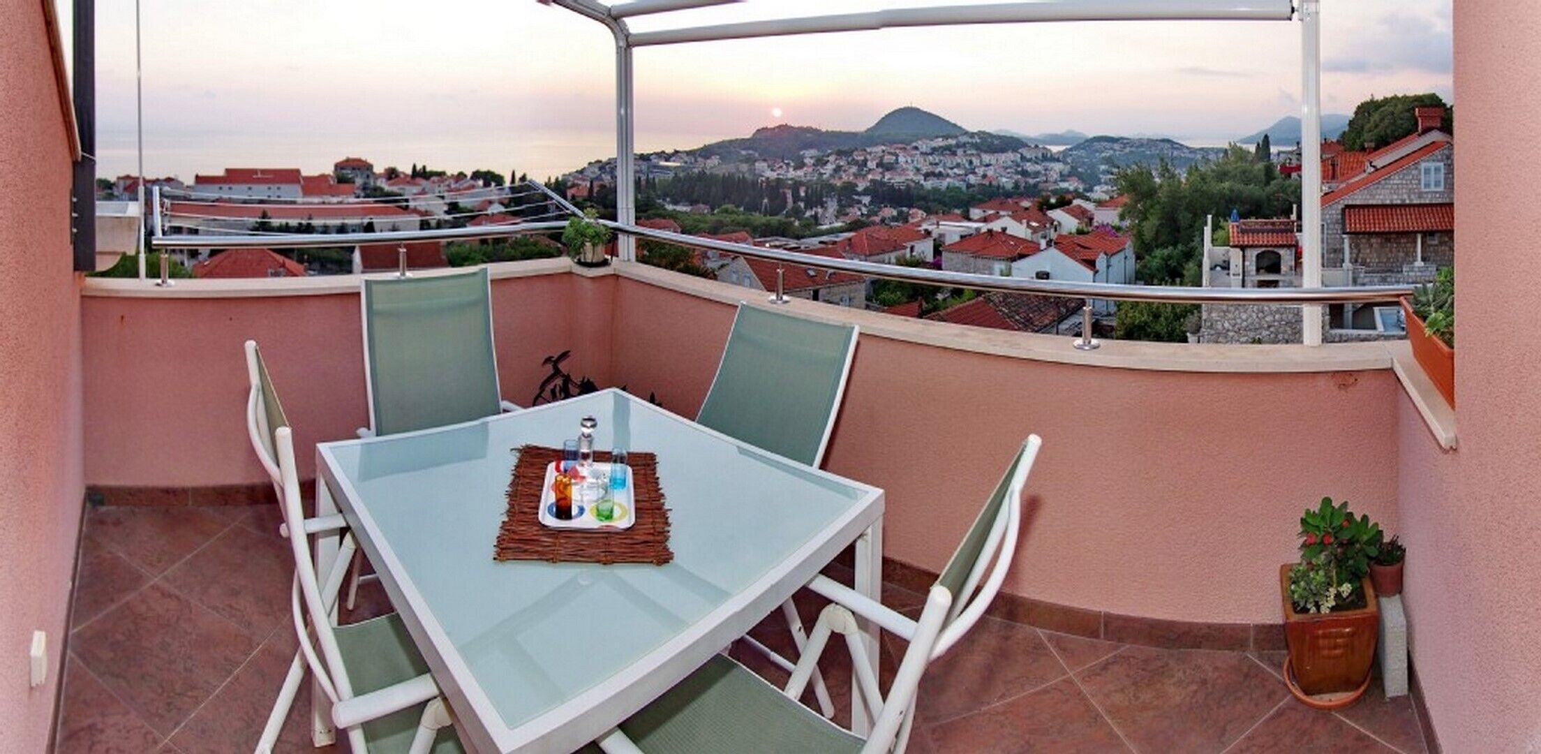 A balcony with a table and chairs, a tray with bottles, and a view of a town and mountains.