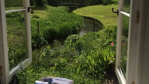 A view through an open window of a garden with a small stream, a wooden bridge, and lush greenery.