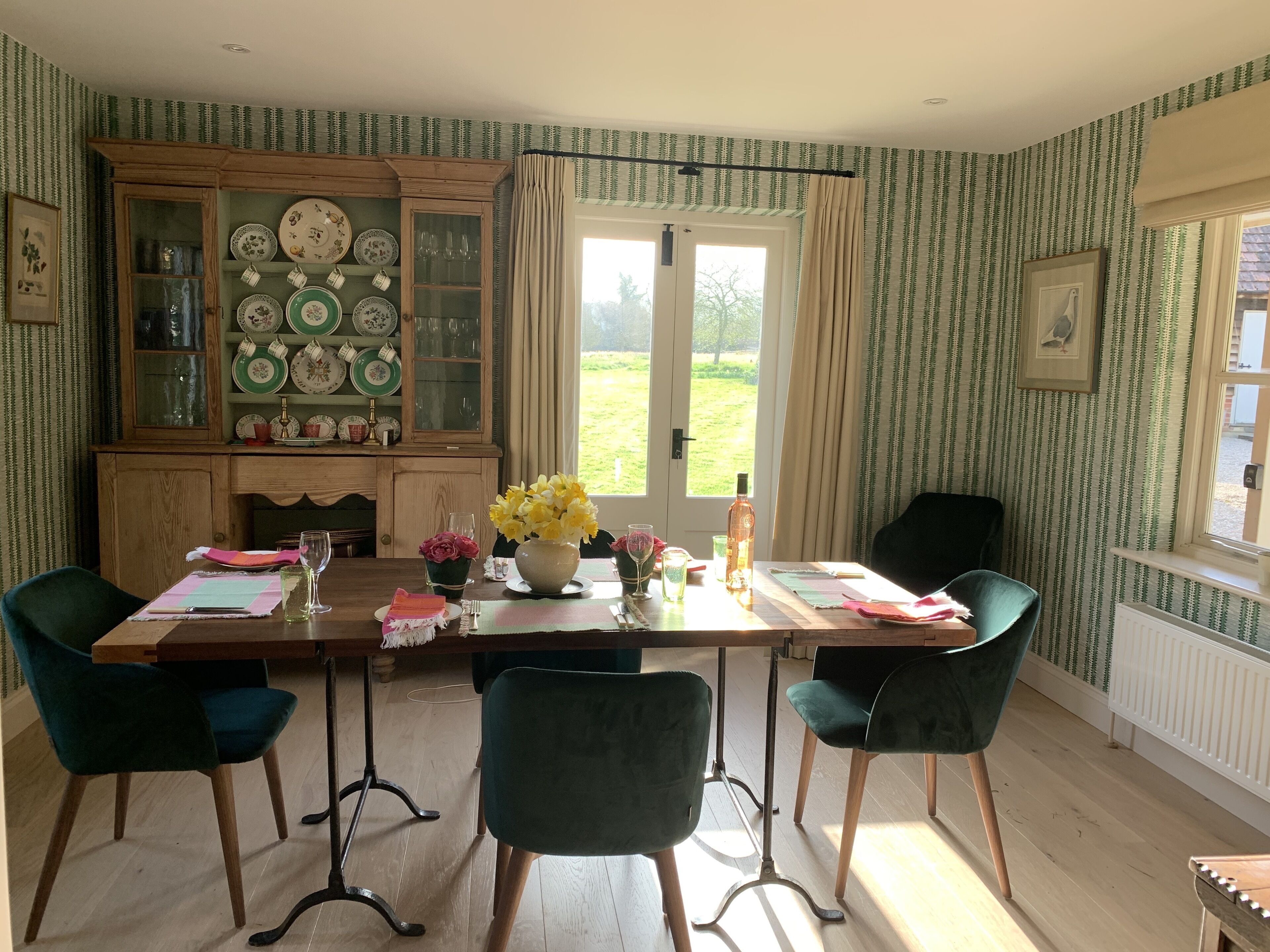 A dining area with a wooden table, four chairs, a cabinet with plates, and a window with curtains.