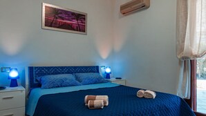 A bedroom with a blue bedspread, two bedside lamps, a wall-mounted air conditioner, and a painting of a sunset.