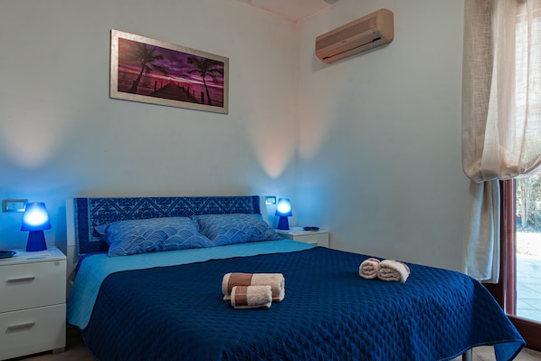 A bedroom with a blue bedspread, two bedside lamps, a wall-mounted air conditioner, and a painting of a sunset. - Welcome to Villa Mirta: Your Tranquil Retreat in Alghero (Alghero)