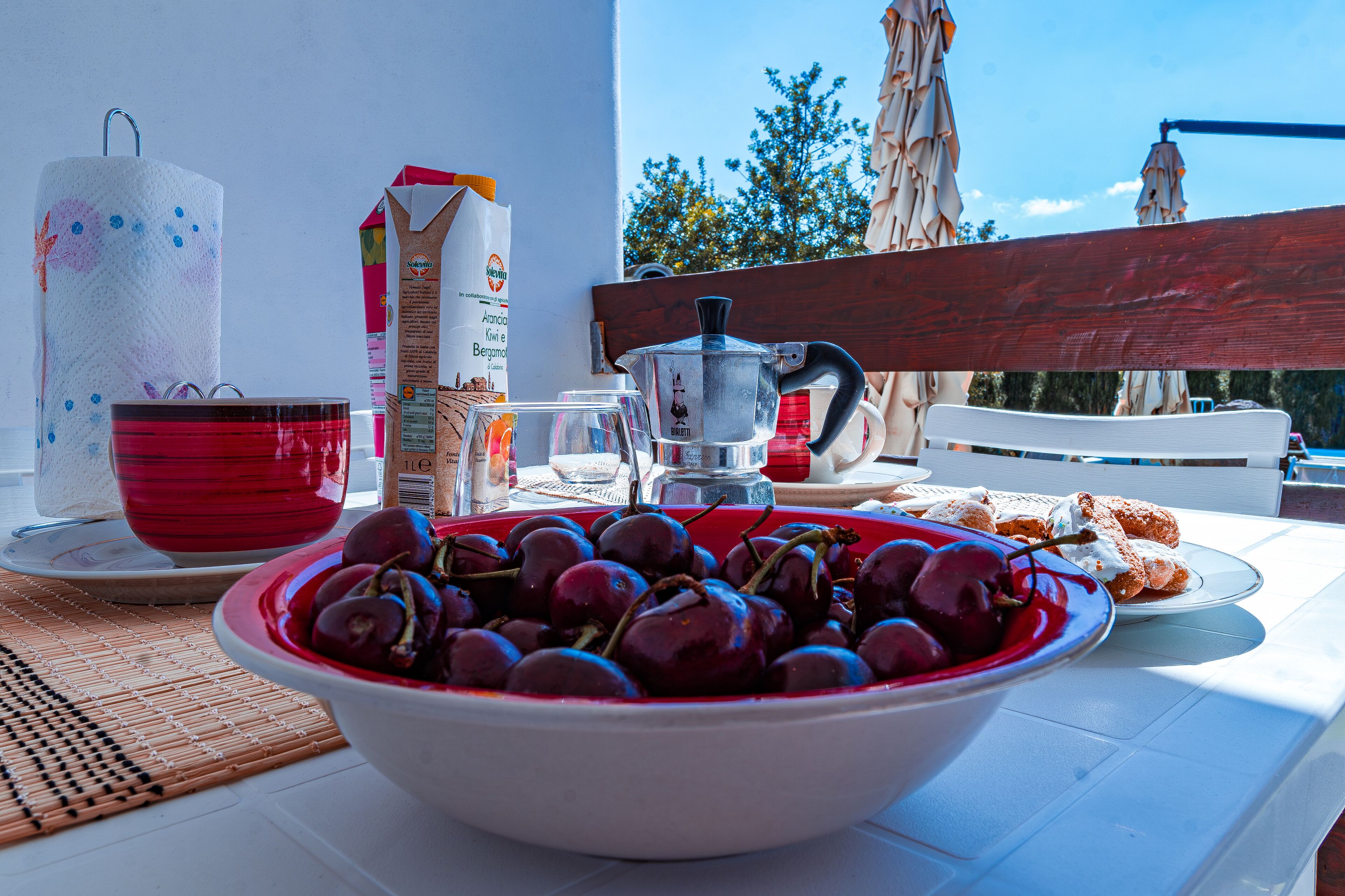 A table with a bowl of cherries, a red cup, a coffee pot, and a carton of milk.