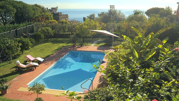 A rectangular swimming pool with a clear blue water surface, surrounded by a brick patio. There are two lounge chairs with umbrellas, and the pool is set against a backdrop of lush greenery and a view of the sea. - Magnificent Villa with private pool and garden (Sanremo)