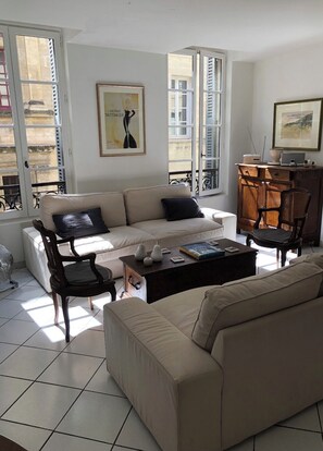 Una sala de estar con un sofá color beige, una mesa de centro de madera y un sillón. Se ve una ventana con contraventanas, un ventilador y un cuadro en la pared.