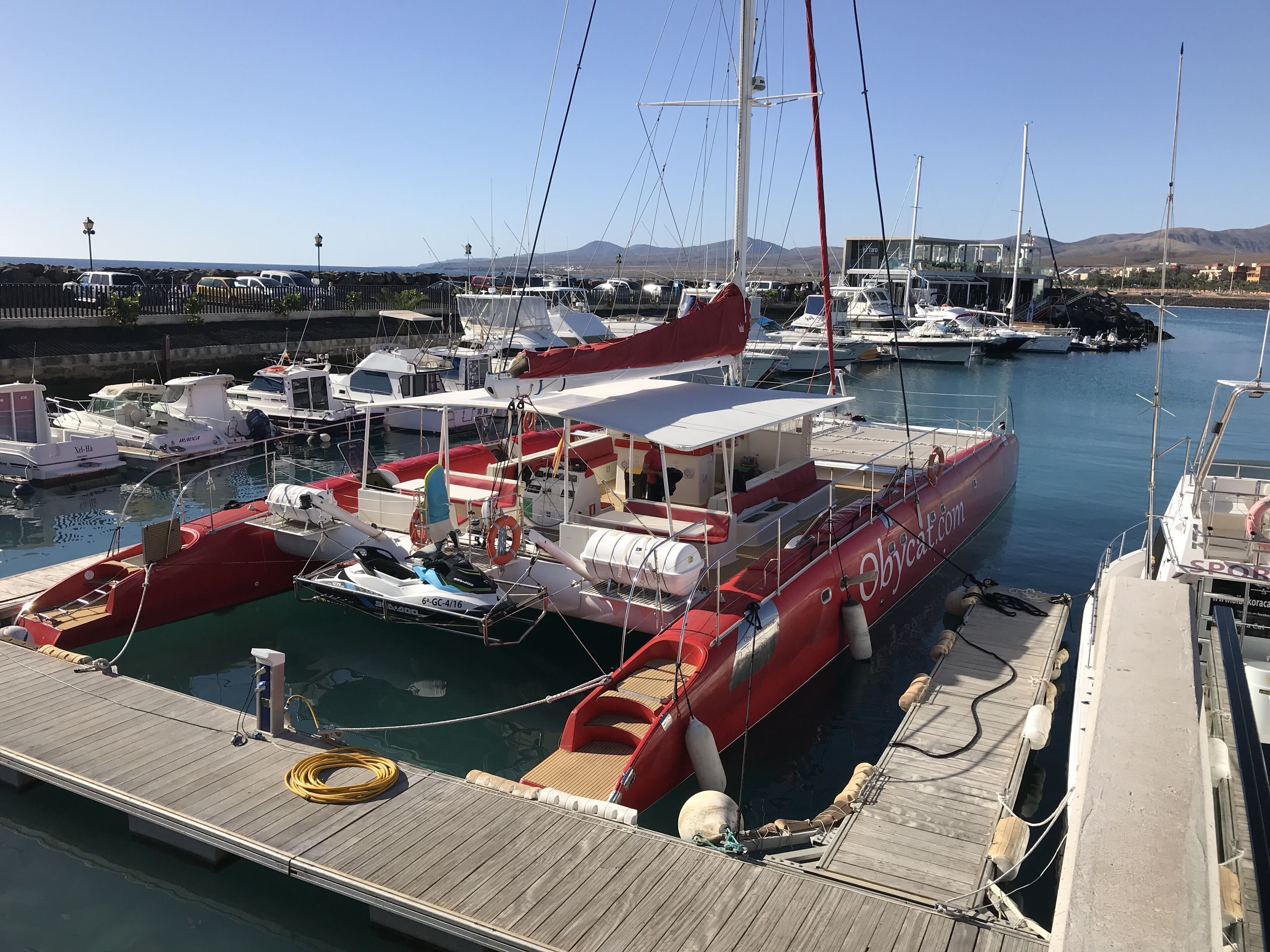 Un port de plaisance où sont amarrés différents types de bateaux, une jetée en bois et, en arrière-plan, un paysage montagneux.