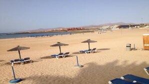Una platja amb gandules i ombrel·les, un cubell de brossa i un edifici al fons.