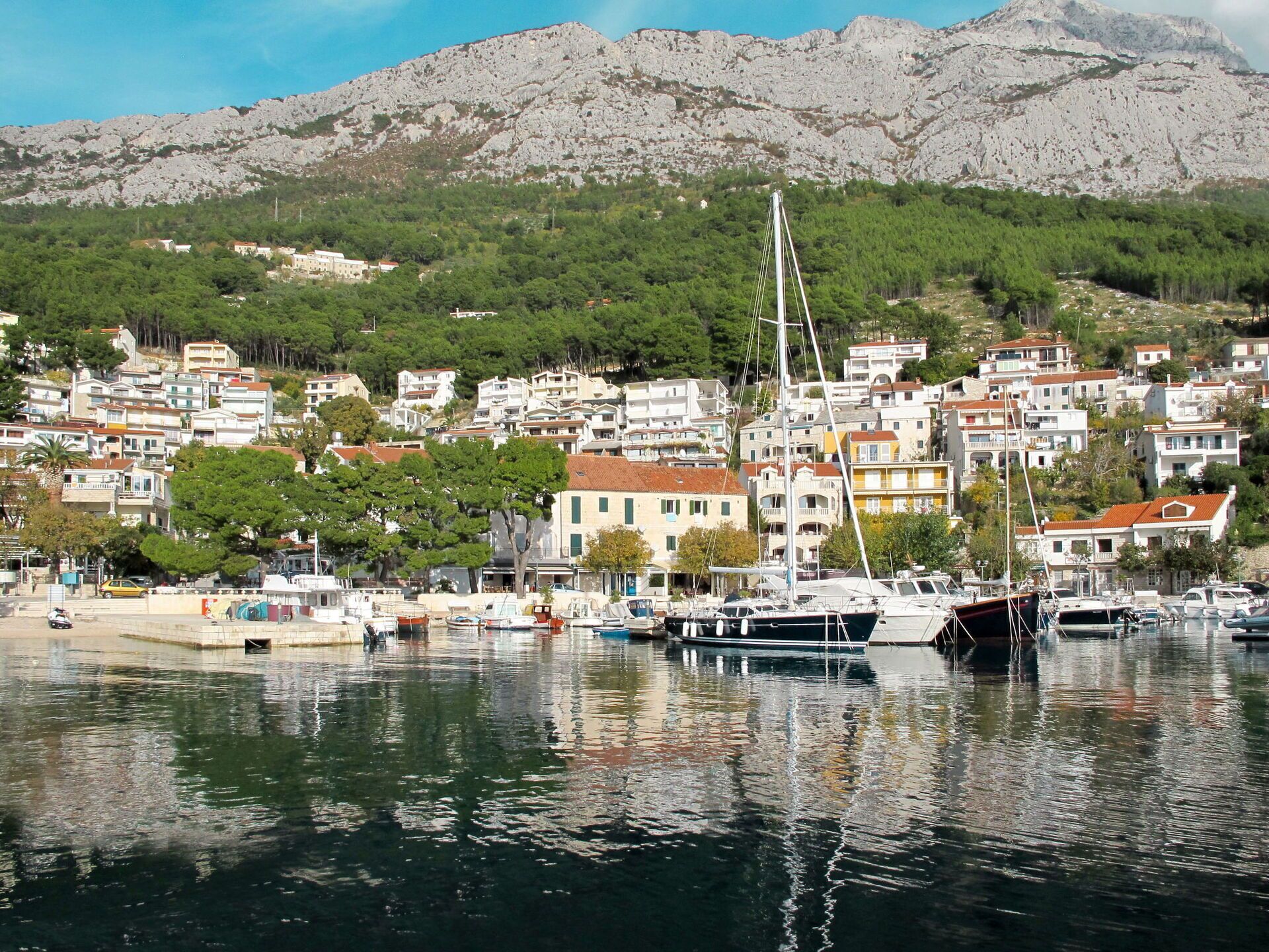 A coastal town with a marina, buildings with red roofs, and a mountainous backdrop.