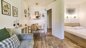 A compact, well-lit room with a wooden floor, a bed, a desk with a chair, a small kitchenette, and a green couch.