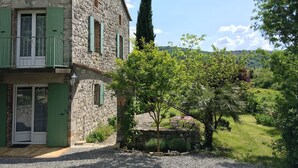 A stone house with a balcony, a door with green shutters, and a garden with a table and chairs.