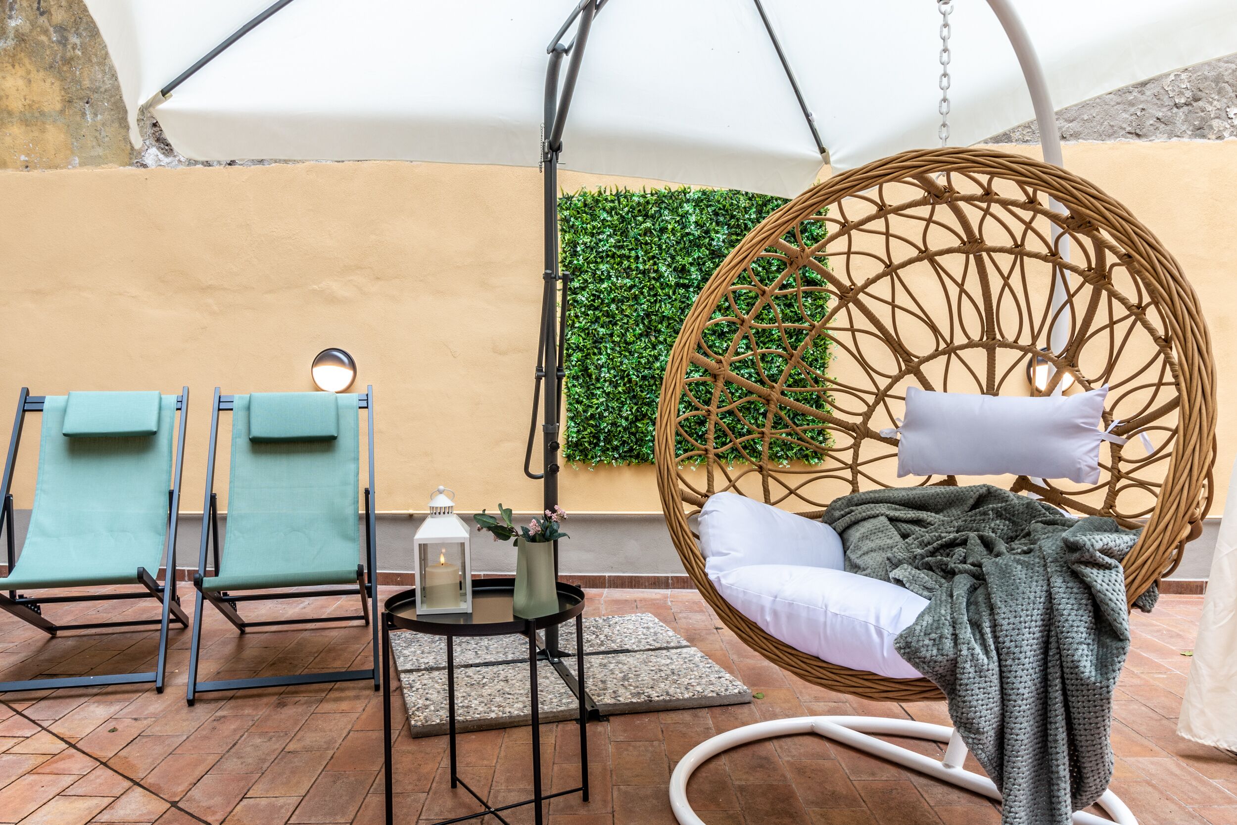 Un patio con dos sillones de descanso, una mecedora de mimbre, una mesita con un farol y una pared verde.