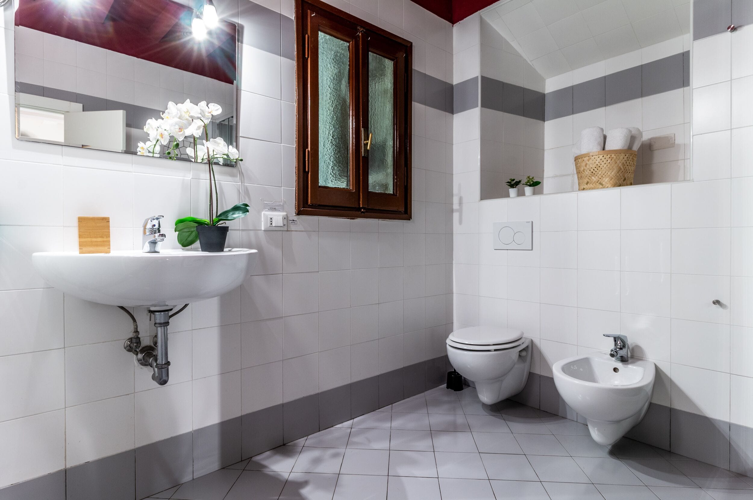 Un baño moderno con un lavamanos blanco, un inodoro y un bidé. Hay un espejo sobre el lavamanos, una ventana con marco de madera y una cesta en la pared.