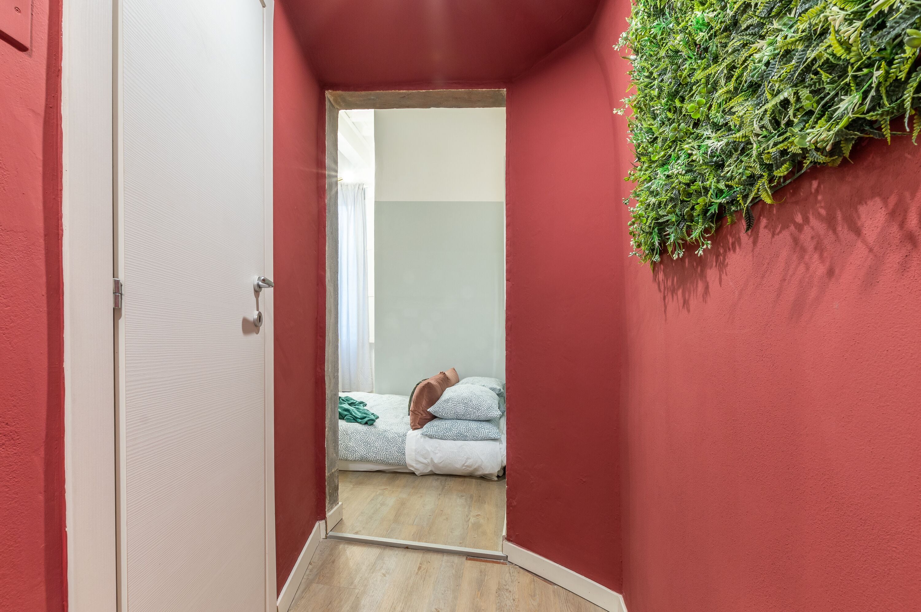 Un corridoio stretto con una parete rossa, una porta bianca e un letto con coperta e cuscini verdi.