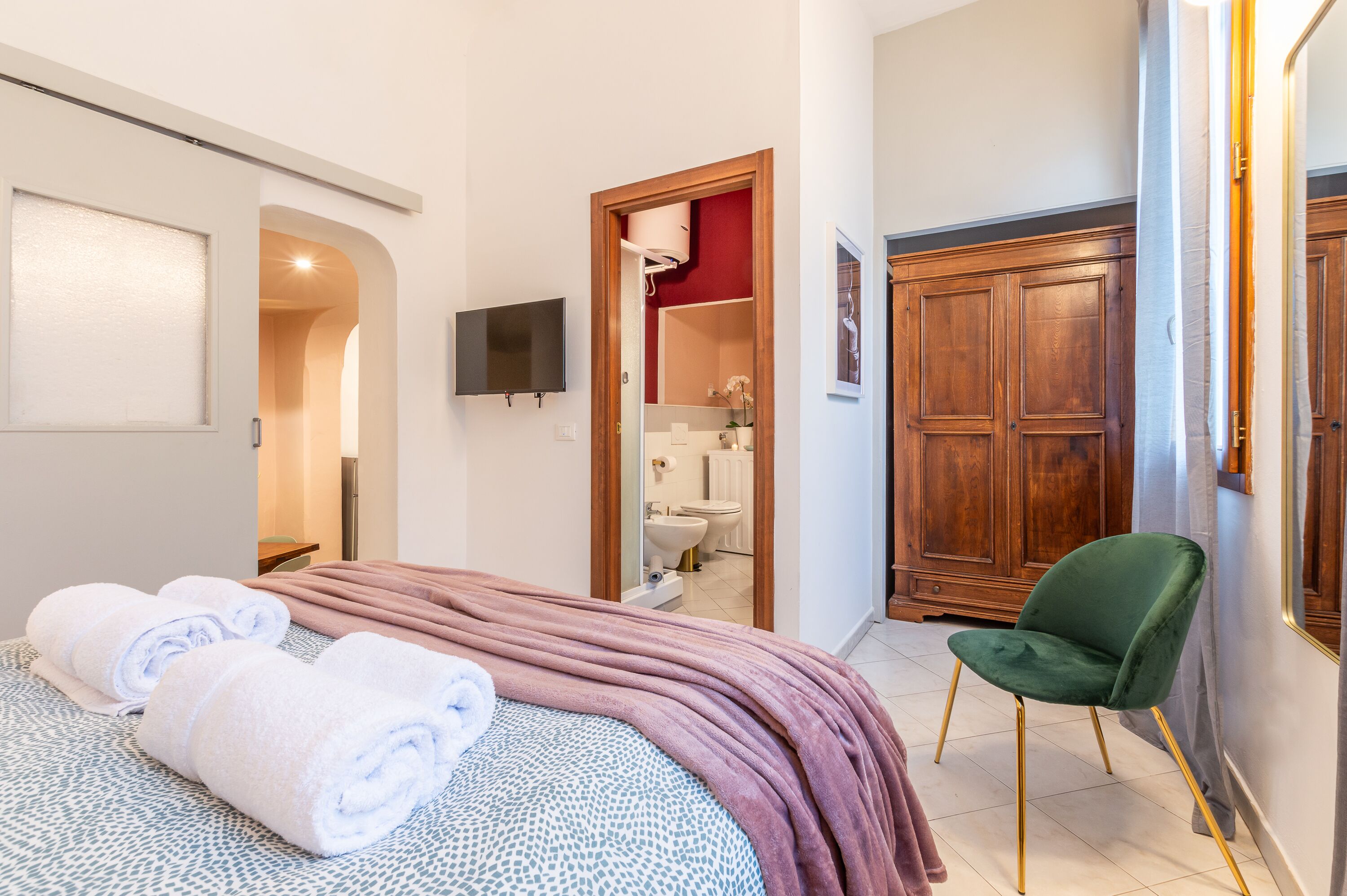 Une chambre à coucher avec un lit, deux serviettes enroulées, un fauteuil vert, une armoire en bois et un téléviseur fixé au mur.