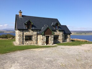 A stone house with a dark roof, a chimney, and a view of a lake.