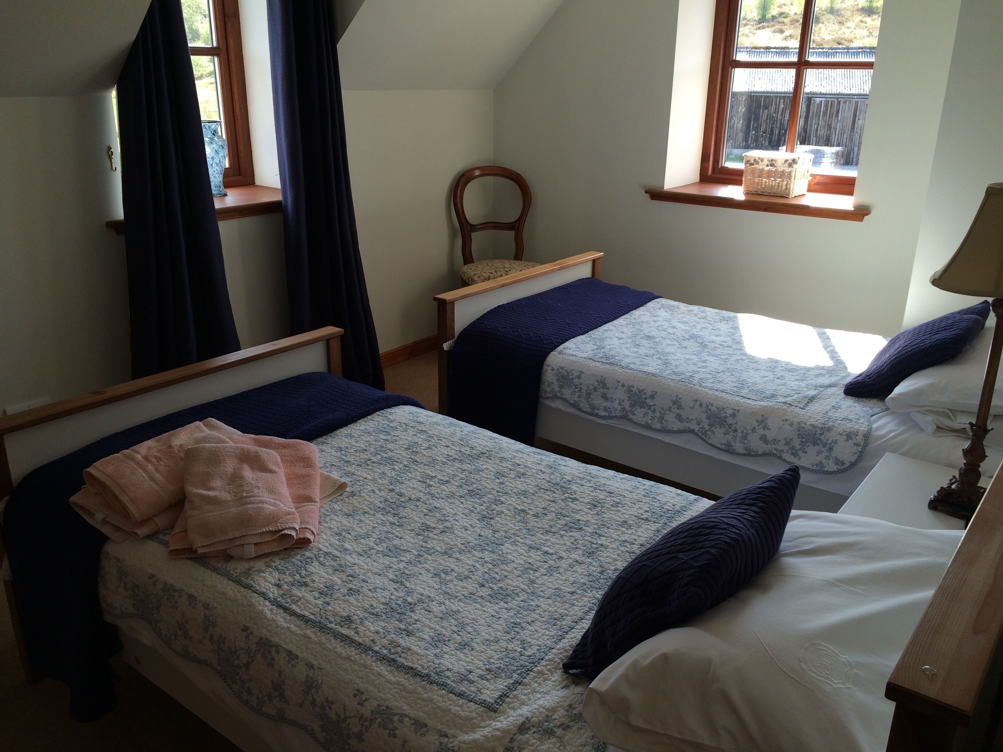 Un dormitorio con dos camas individuales, una silla de madera, una ventana con vistas al exterior y una pequeña mesa con una cesta.