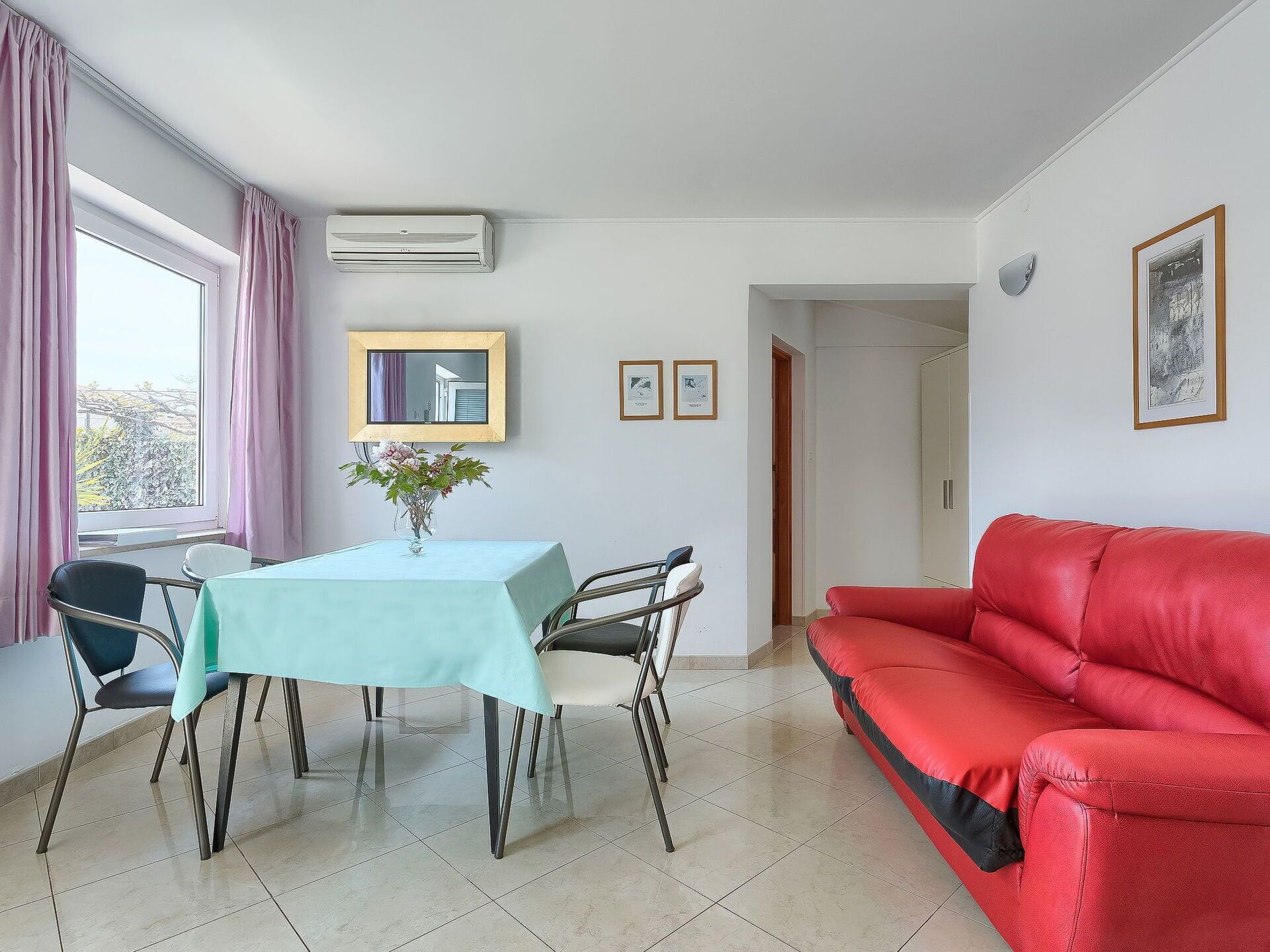 A room with a dining table, chairs, a red sofa, a window with curtains, and a wall-mounted air conditioner.