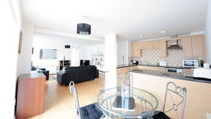 Eine moderne Küche mit Kochinsel, ein Essbereich mit Glastisch und ein Wohnzimmer mit schwarzem Sofa.